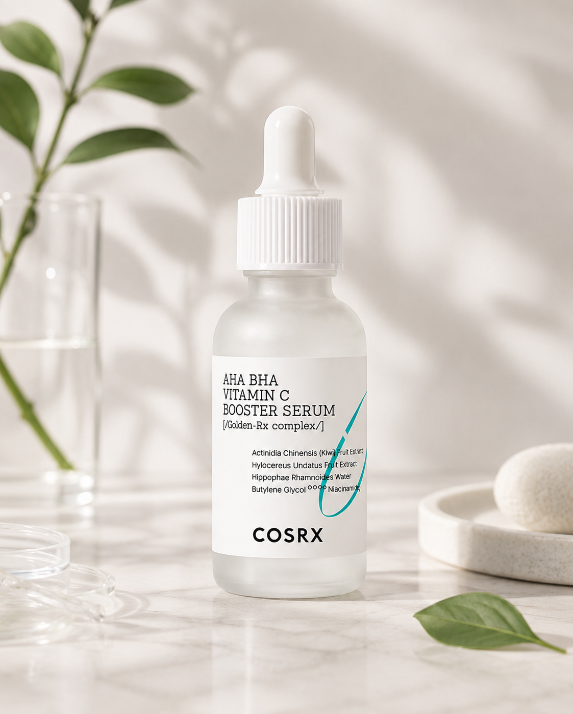 COSRX AHA BHA Vitamin C Booster Serum in a clear dropper bottle with a white cap, set on a marble surface with soft leaf shadows, a glass vase, and a soap dish in the background.