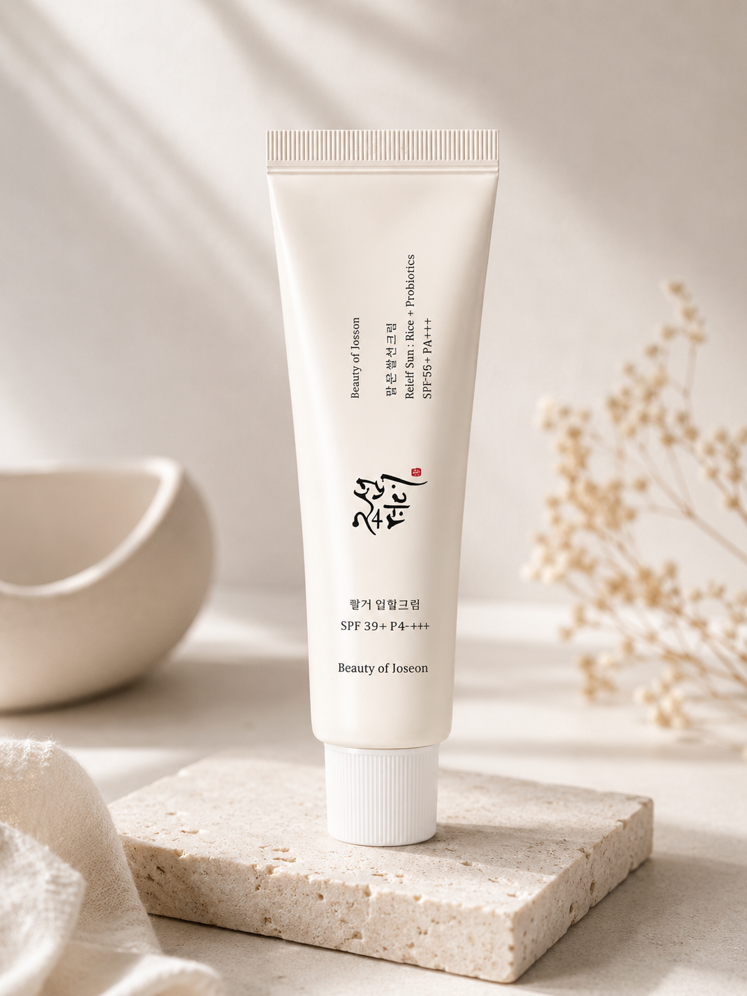 Creamy off-white sunscreen tube with rice and probiotics SPF50+ PA++++ by Beauty of Joseon, standing upright on stone slab with dried flowers and ceramic bowl in soft-lit background.
