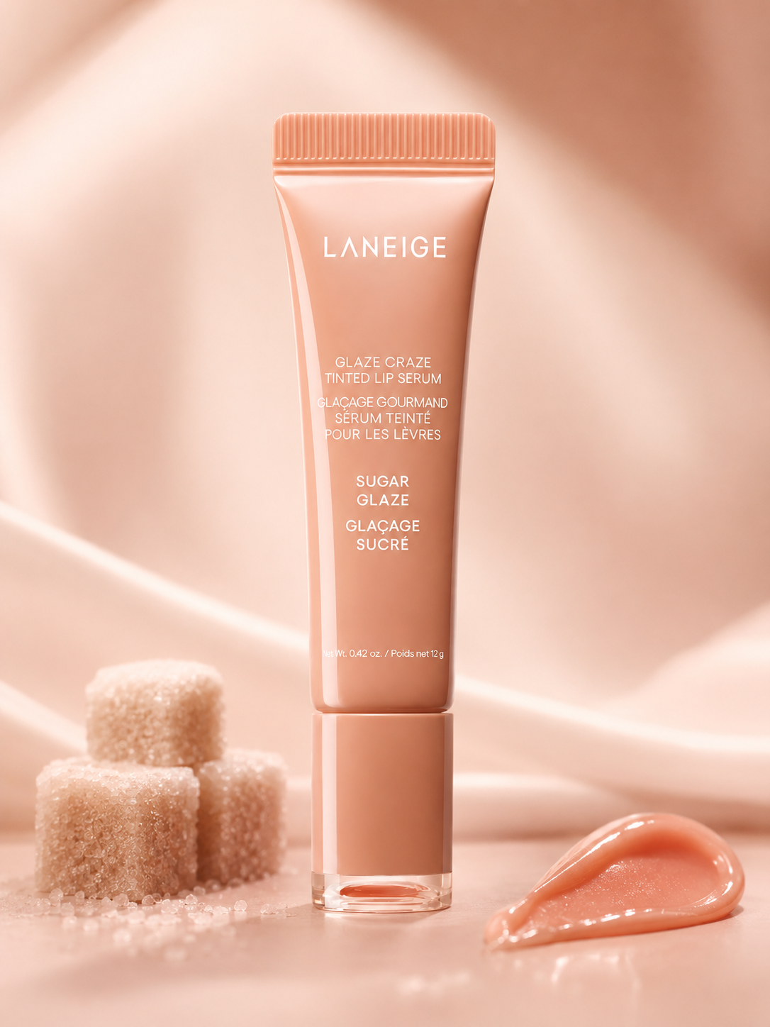 A tube of Laneige Glaze Craze Tinted Lip Serum in a soft peach shade is displayed among sparkling sugar cubes and a glossy lip serum droplet on a satin surface.