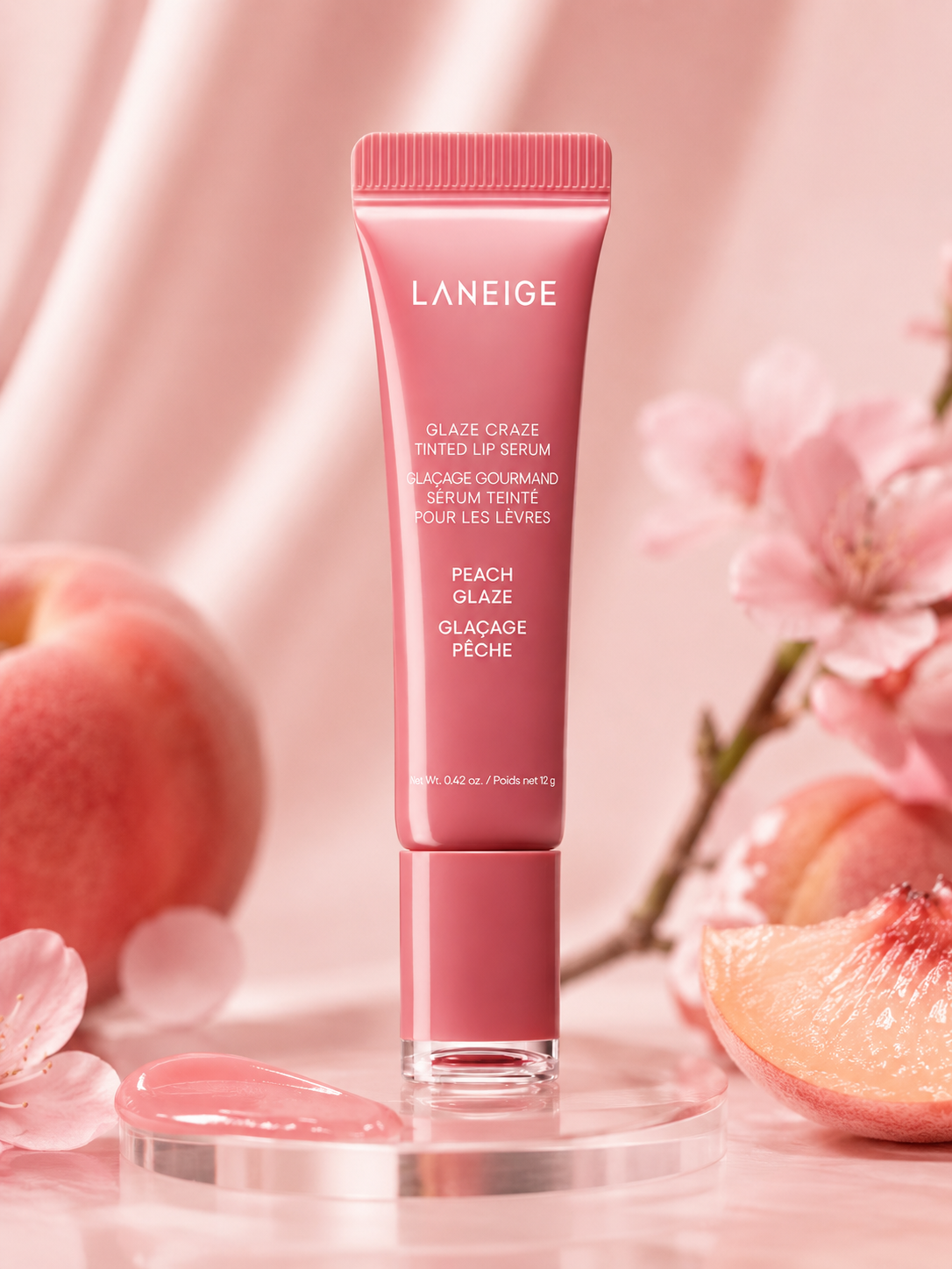 A tube of Laneige Glaze Craze Tinted Lip Serum in Peach Glaze is positioned among fresh peach slices and pink flowers, showcasing its vibrant packaging and creamy texture.